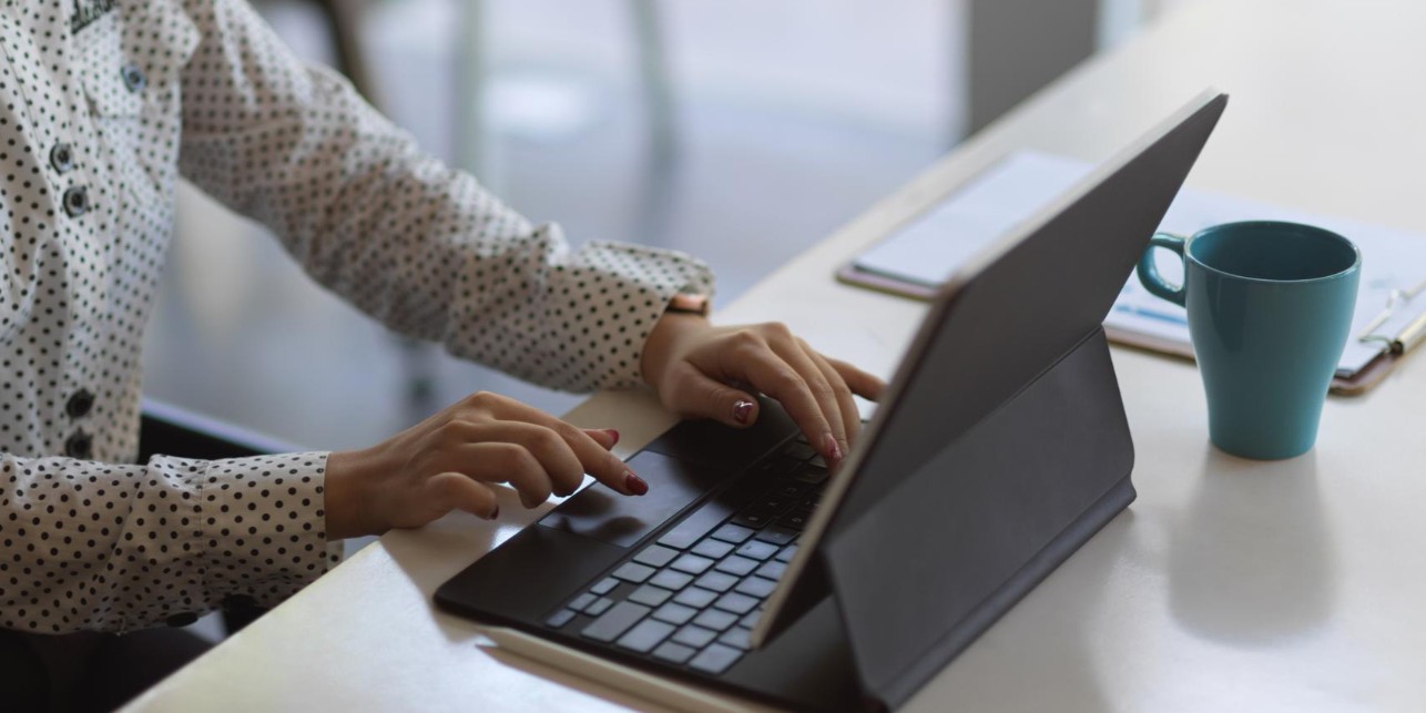 Osoba pisząca na klawiaturze laptopa, obok laptopa stoi kubek i podkładka na dokumenty