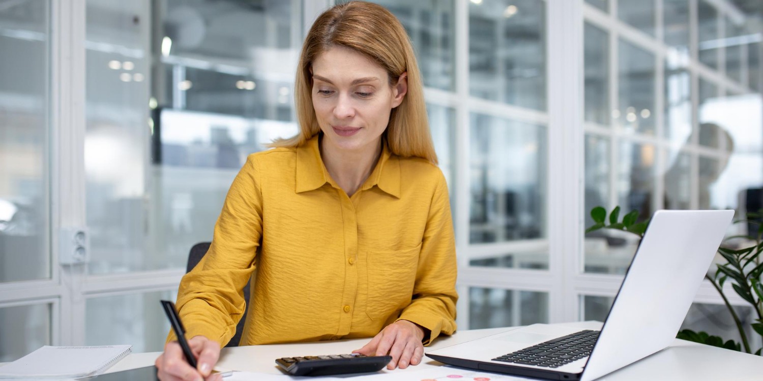 Kobieta licząca coś na kalkulatorze, obok kalkulatora leży laptop
