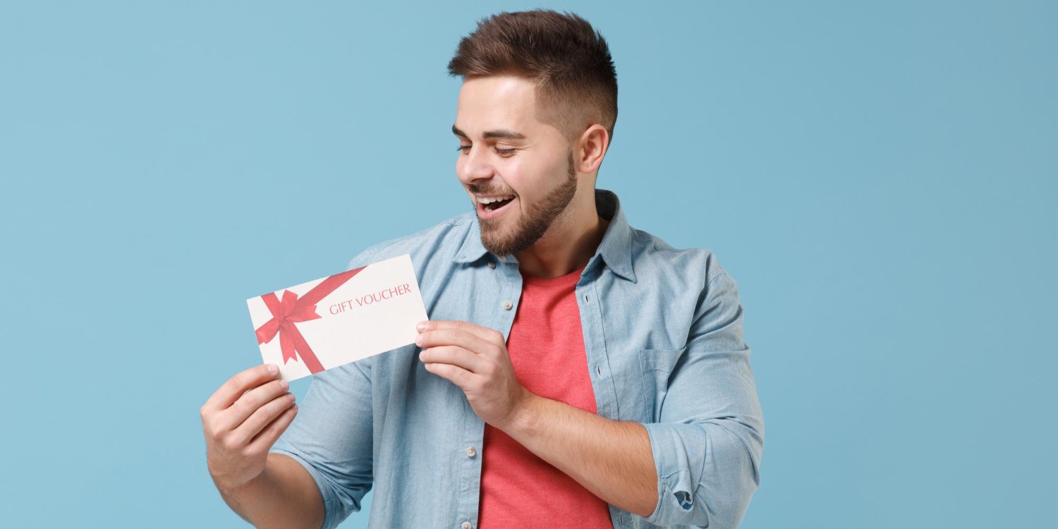 Uśmiechnięty, młody, brodaty mężczyzna po dwudziestce w luźnej koszuli. Trzyma kartę podarunkową.