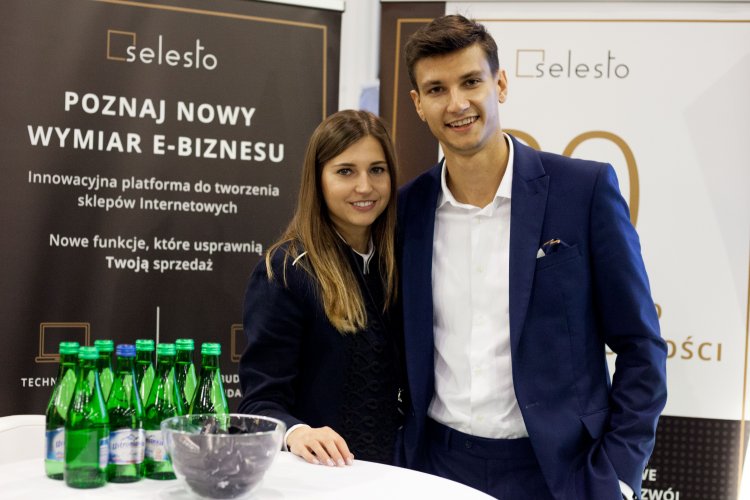 Dwie osoby stojące obok siebie przy stoliku z butelkami wody; osoby pozują na tle ścianek reklamujących firmę Selesto