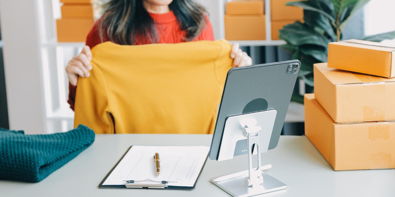 Kobieta trzymająca sweter, na biurku tablet, kartka na podkładce i długopis, obok kilka paczek