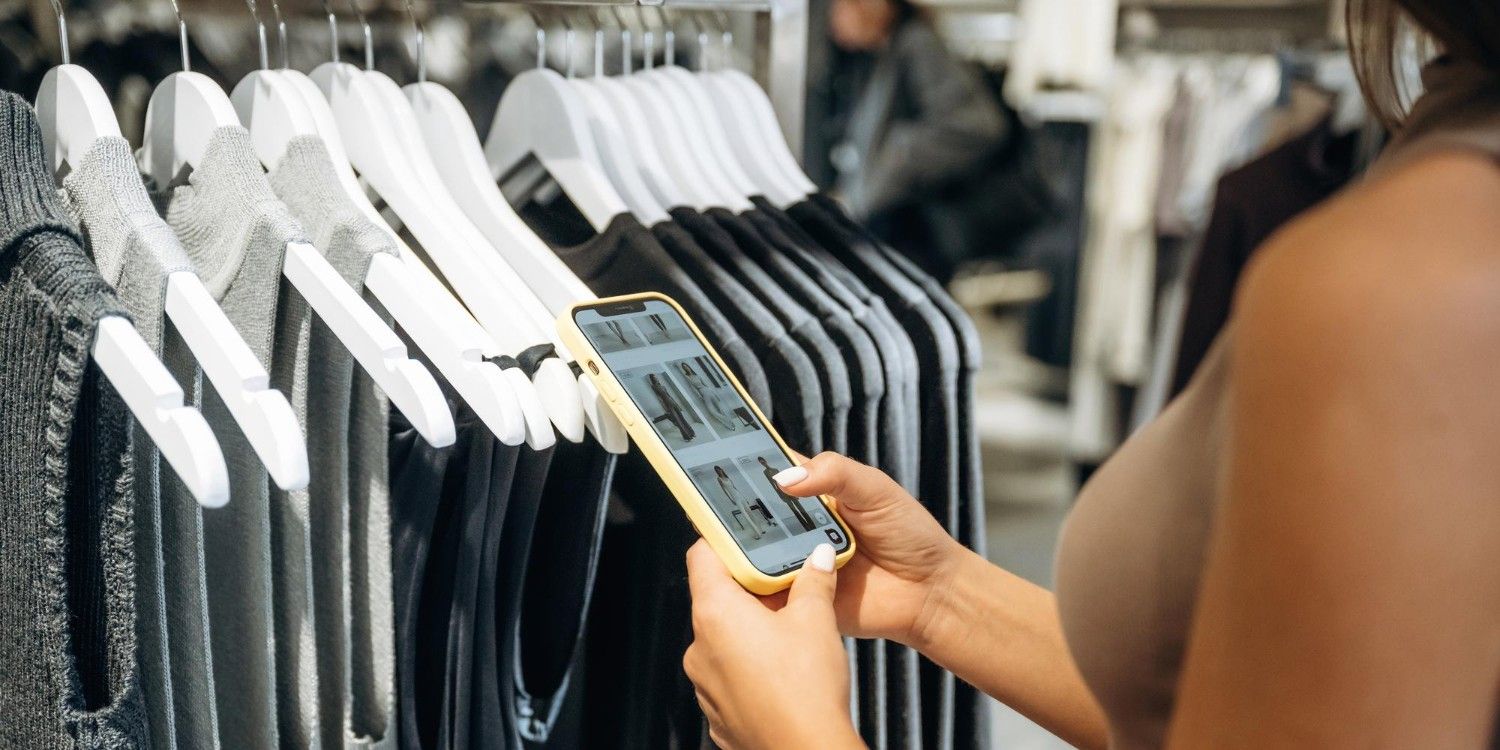 Kobieta przeglądająca ubrania w smartfonie, przed nią wiszące na wieszakach ubrania